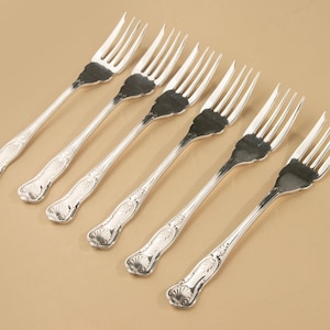 Könnte beinhalten: Ein Set aus sechs silberfarbenen Metallgabeln, die in einer diagonalen Reihe angeordnet sind. Jede Gabel hat einen Kopf mit vier Zinken und einen Griff, der mit einem Muschelmotiv am Ende und feinen Verzierungen am Stiel versehen ist.