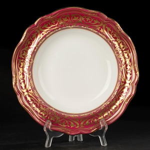 Könnte beinhalten: Eine weiße Porzellanschale mit einem tief crimsonroten Rand und einem goldenen, vergoldeten Blumenmuster. Die Schale steht auf einem weißen Ständer.