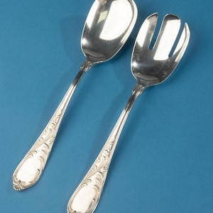 Pode incluir: Um par de utensílios de servir salada em tom prateado, composto por uma colher sólida e um garfo com ranhuras. Ambas as peças apresentam um acabamento polido e um padrão ornamental em relevo ao longo dos cabos, terminando numa base arredondada.