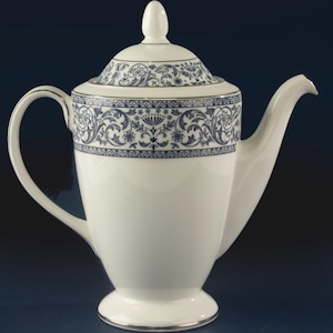 May include: A white porcelain coffee pot featuring a dark blue floral and scrollwork pattern around the upper body and lid. The pot has a curved handle, a pointed spout, a pedestal base, and a matching lid with a small finial handle on top.
