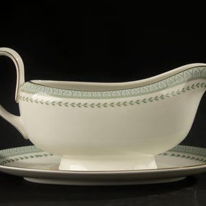 May include: A cream-colored gravy boat with an attached oval underplate, featuring a sage green band with a repeating leaf and fan pattern along the rim and a thin gold trim on the edges, handle, and base of the ceramic serving vessel.