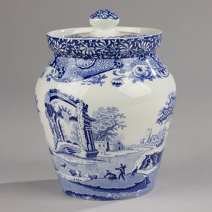 Puede incluir: Un tarro de cerámica blanco con tapa, con una escena pastoral azul y blanca. El tarro tiene una forma redondeada con un patrón floral detallado alrededor del borde y en el pomo de la tapa. El diseño representa un paisaje con figuras y edificios.