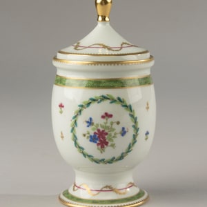 Könnte beinhalten: Ein weißes Porzellangefäß mit Deckel, goldenem Knauf, grünem Punktband und einem zentralen Blumenkranzmotiv mit roten und blauen Blüten. Der Boden und der Deckelrand sind mit Goldverzierungen und feinen roten Linien dezent gestaltet.