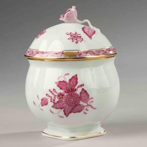 Pode incluir: Um pote com tampa de porcelana branca com um design floral. O pote apresenta um corpo arredondado, uma tampa com uma pega de flor rosa e padrões florais rosa. As bordas douradas acentuam a borda e a base, adicionando um toque de elegância à peça decorativa.