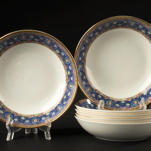 May include: A set of white ceramic bowls featuring a wide rim decorated with a blue, pink, and gold patterned border. The bowls have a smooth, glossy finish and a gold-toned edge. One bowl is displayed upright on a stand, while others are stacked.