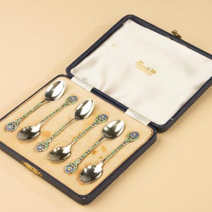 May include: A set of five silver-toned spoons with blue and green enamel floral detailing on the handles, presented in a dark blue hinged case with a white satin interior lining featuring gold text that reads Tostrup Oslo Norway.