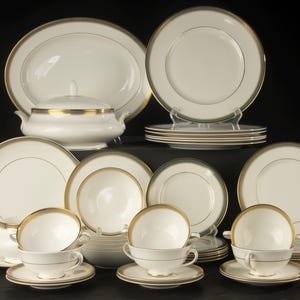 Può includere: Un set di stoviglie in porcellana color bianco sporco con bordi dorati, composto da piatti, ciotole e tazze. La collezione include una zuppiera con coperchio, piatti di varie dimensioni e tazze da tè con piattini abbinati dal design uniforme.