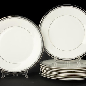 Op de afbeelding: Een set witte keramische dinerborden met een dunne zwarte rand en een verfijnde kralentextuur. Eén bord staat rechtop op een doorzichtige kunststof standaard, terwijl een stapel van meerdere bijpassende borden ernaast is geplaatst.