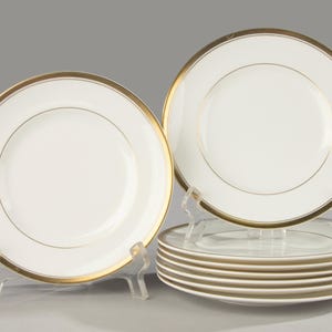 Può includere: Un set di piatti in ceramica bianca con un ampio bordo in oro metallizzato e una sottile linea circolare dorata vicino al centro. Un piatto è esposto in verticale su un supporto trasparente, mentre gli altri sono impilati orizzontalmente.