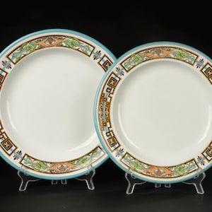 Op de afbeelding: Twee witte keramische borden met een turquoise rand en decoratieve versieringen. De versieringen hebben geometrische patronen in bruin, zwart en groen. De borden worden gepresenteerd op transparante standaards tegen een zwarte achtergrond.