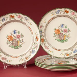 Pode incluir: Um conjunto de cinco pratos de jantar em cerâmica de cor creme com um motivo floral central de flores cor de laranja, rosa e azul. Os pratos têm uma borda recortada com um rebordo verde e uma borda geométrica castanha central.