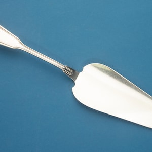 May include: A silver-toned cake server featuring a polished, triangular blade and a slender handle with a rounded, heart-shaped decorative detail at the end. The metal surface reflects light, showing a smooth, metallic finish throughout the utensil.