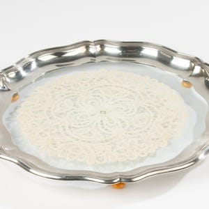 May include: A silver serving tray with a decorative, scalloped edge. It features a cream-coloured, circular lace doily. The tray is supported by small, amber-coloured feet.