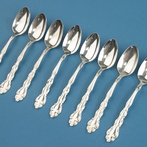 Puede incluir: Un juego de ocho cucharas de metal color plata dispuestas en una fila diagonal. Cada cuchara presenta un cuenco ovalado pulido y un mango decorativo con volutas en relieve y detalles florales a lo largo de los bordes y la base.
