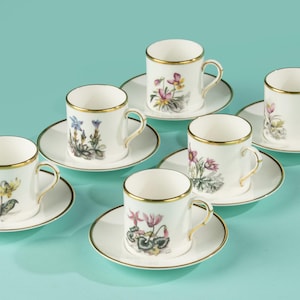 Puede incluir: Seis tazas de espresso de porcelana blanca con bordes y asas doradas. Cada taza presenta un diseño floral diferente en tonos amarillos, rosas, morados y azules. El juego está dispuesto sobre una superficie azul claro.