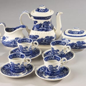 Op de afbeelding: Een keramische theeset met een crèmekleurige basis en een blauw patroon in willow-stijl. De set bevat een theepot met deksel, een melkkan, een suikerpot met deksel en vier theekopjes met bijpassende schoteltjes, voorzien van gedetailleerde landschappen.