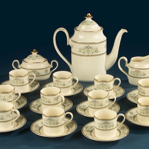 May include: A porcelain tea set featuring a large teapot, a lidded sugar bowl, a creamer, and eight cups with matching saucers. Each piece is cream-coloured with a decorative band of blue and green floral patterns and gold-toned trim accents.