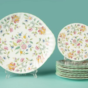 May include: A collection of floral patterned plates. The set includes a large square plate and a stack of smaller round plates. The plates feature a white background with colourful flowers in pink, yellow, and blue, with green accents. The plates are displayed on clear stands.