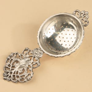 May include: A silver-toned metal tea strainer featuring a deep, perforated bowl and an ornate, filigree-style handle with intricate scrollwork and leaf-like patterns. The rim of the bowl is decorated with a repeating notched edge design.