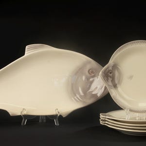 Peut inclure: Un grand plat de service en céramique blanc cassé en forme de poisson, accompagné d'un ensemble assorti de cinq petites assiettes rondes en forme de poisson. Chaque pièce présente des détails de nageoires et d'écailles en relief.