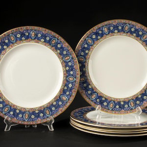 May include: A set of five round white ceramic dinner plates featuring a wide decorative border. The border displays a dark blue background with a repeating pattern of geometric shapes in shades of pink, light blue, and gold, finished with a gold rim.