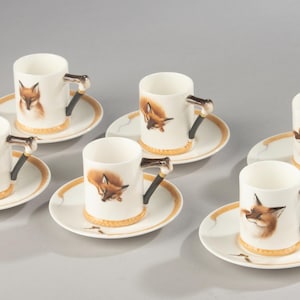 Könnte beinhalten: Ein Set aus fünf weißen Espresso-Tassen aus Keramik mit passenden Untertassen, jede mit einem gemalten Fuchskopf auf der Tasse und einem Henkel in Form eines Fuchsschwanzes. Die Untertassen haben einen goldfarbenen Rand und ein Fuchsschwanz-Detail.