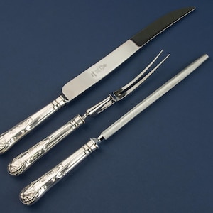 May include: A silver carving set on a dark blue background. The set includes a carving knife, a carving fork, and a sharpening steel. The handles have a decorative floral design. The knife blade is approximately 12 inches long.
