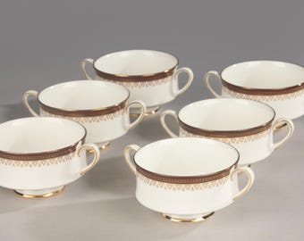 May include: A set of five white ceramic soup bowls, each featuring two side handles and a decorative band around the rim. The band consists of a dark brown border with a repeating gold geometric pattern underneath, and a gold-trimmed base.