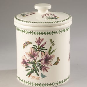 Op de afbeelding: Een cilindrische keramische voorraadpot met bijpassend deksel en knopgreep. Het crèmekleurige oppervlak is voorzien van een botanische illustratie van roze Rhododendron Liliflorum bloemen, groene bladeren, vlinders en een libel.
