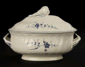 May include: Cream-colored ceramic serving dish with a lid, featuring blue floral designs and a bird-shaped handle. The dish has two handles and a scalloped edge. The lid has a bird-shaped handle and blue floral accents.
