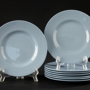 Peut inclure: Un ensemble de huit assiettes en céramique bleu clair, chacune présentant une finition lisse et brillante avec une série de rainures circulaires concentriques en relief sur le bord extérieur. Une assiette est posée sur un support transparent.