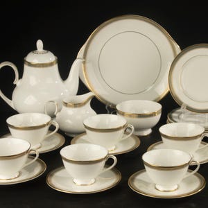 May include: A white porcelain tea set featuring a teapot, creamer, sugar bowl, and six teacups with matching saucers. Each piece is decorated with a thin gold rim and a subtle grey patterned border. A large serving platter sits behind the set.