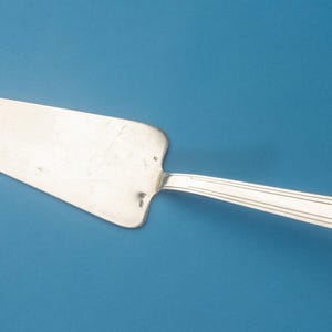 Pode incluir: Um servidor de bolo de metal prateado com uma lâmina triangular larga e plana, e um cabo longo e fino com um design linear simples em relevo. A superfície do metal apresenta um acabamento polido com sinais de desgaste e riscos leves.