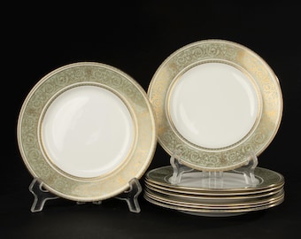 May include: A set of fine china plates with a white center and a decorative border in gold and olive green. The plates are arranged on clear plastic stands against a black background. The set includes several plates stacked together.