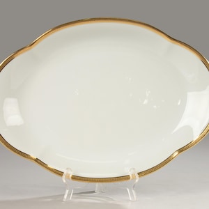 Op de afbeelding: Een ovale, witte porseleinen serveerschaal met een geschulpte rand en een gouden rand. De gouden rand heeft een herhalend geometrisch patroon. De schaal wordt gepresenteerd op een transparante plastic standaard.