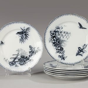 Könnte beinhalten: Set aus acht weißen Porzellantellern mit schwarzen und blauen Blumen- und Vogelmotiven. Die Teller haben einen gewellten Rand und ein zartes Blumenmuster.