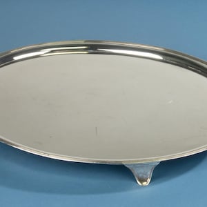 Peut inclure: Un plateau de service ovale de couleur argentée polie avec une surface lisse et réfléchissante et un bord surélevé et rainuré. Le plateau est soutenu par trois pieds courts et incurvés qui surélèvent légèrement la base.