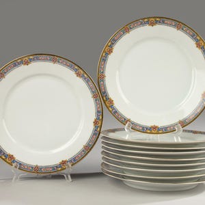 Op de afbeelding: Een set witte porseleinen borden met een decoratieve rand. De rand heeft een blauw, rood en goudkleurig bloemmotief. De borden hebben een gouden rand. De afbeelding toont twee borden op standaards en een stapel borden.