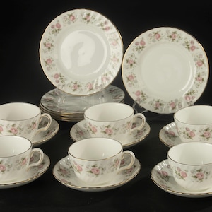 May include: A set of white porcelain teacups and saucers featuring a delicate pink rose floral pattern with green leaves and gold trim. The collection includes multiple matching cups, saucers, and small plates arranged on a dark surface.