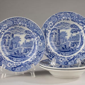 May include: A set of four white ceramic bowls featuring a blue transferware pattern. The design depicts a landscape with classical ruins, trees, and figures, surrounded by a wide, intricate floral and geometric border on the rim of each bowl.