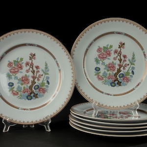 Op de afbeelding: Een set witte porseleinen borden met een centraal bloemmotief van roze, blauwe en groene bloesems op bruine takken. De rand is versierd met een metallic gouden band en een herhalend bruin geometrisch patroon langs de rand.
