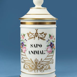 May include: A white ceramic apothecary jar with a lid and gold accents. The jar features the text "SAPO ANIMAL:" in black, surrounded by floral and gold scrollwork designs. The lid has a gold knob.