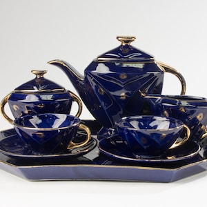 May include: A navy blue tea set with gold trim, comprising a teapot, sugar bowl, creamer, two teacups and saucers, and a serving tray. The set features a geometric pattern with gold detailing. The tray is a hexagonal shape.