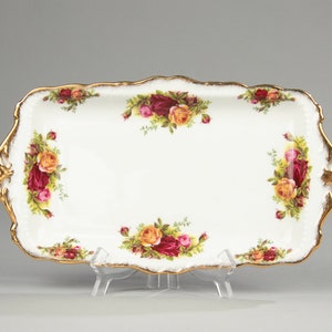 Puede incluir: Una bandeja rectangular de cerámica blanca con un borde dorado festoneado. La bandeja está decorada con racimos de rosas rojas, rosas y amarillas. La bandeja está sostenida por un soporte de plástico transparente.