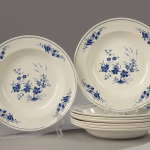 Op de afbeelding: Een set witte keramische kommen met een blauw bloemmotief. De kommen hebben een brede rand met een blauwe rand en zijn op een transparante standaard gestapeld. Het bloemmotief herhaalt zich rond de rand en in het midden van de kommen. Elke kom meet ongeveer 25 cm in diameter.