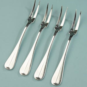 Puede incluir: Cuatro tenedores plateados con tres púas cada uno, dispuestos en diagonal sobre un fondo azul claro. Los tenedores tienen un diseño clásico con mangos alargados y un acabado pulido, adecuados para servir aperitivos o postres. Miden aproximadamente 15 cm.