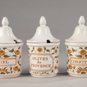 Può includere: Tre barattoli di ceramica bianca con coperchi, adornati con motivi floreali arancioni e verdi. I barattoli hanno etichette con le scritte "MIEL", "OLIVES de PROVENCE" e "CONFITURE".