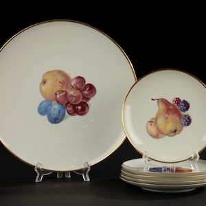 May include: A set of white porcelain plates with gold rims. The plates feature colorful fruit illustrations, including apples, plums, grapes, and pears. The plates are displayed on clear stands against a dark background.