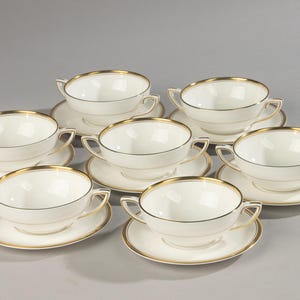 Può includere: Un set di sei tazze da brodo in porcellana bianca con piattini, ciascuna dotata di due piccoli manici e un sottile bordo dorato sul bordo superiore della tazza e sul bordo esterno del piattino. Design semplice con finitura liscia e lucida.
