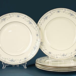 May include: A set of six round ceramic dinner plates featuring an off-white centre and a decorative border of blue floral patterns and small leaves. Each plate is finished with a thin silver metallic rim along the outer edge.